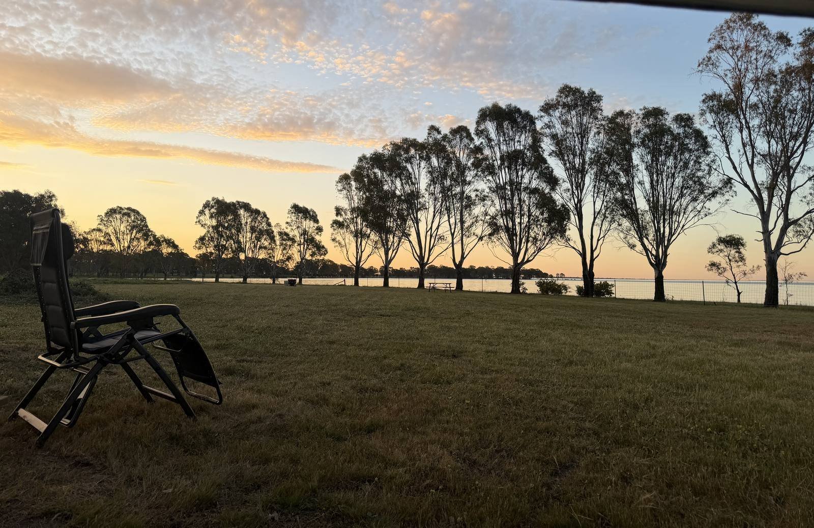 An empty camp chair facing the lake at first light, gum trees along the far bank